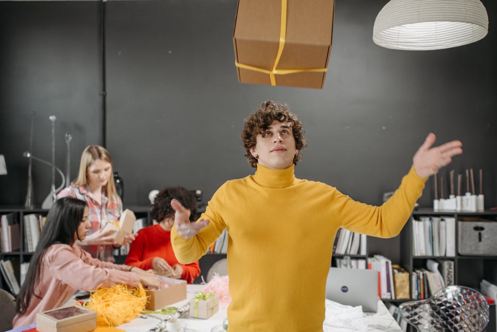 A young man in a yellow sweater tossing a gift box in a creative office setting with colleagues.