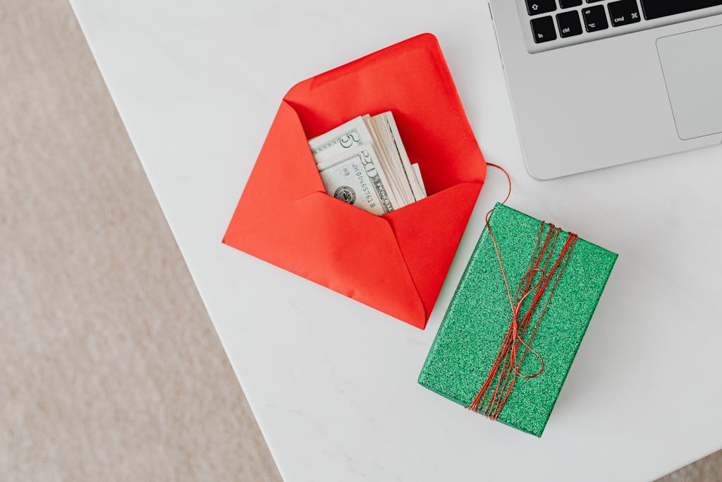 A green gift box and red envelope with cash on a desk beside a laptop, emphasizing holiday or financial themes.
