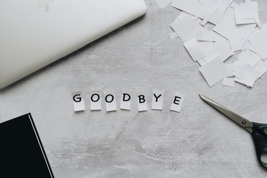 Artistic flat lay featuring the word 'GOODBYE' crafted from paper cutouts with scissors and a laptop.