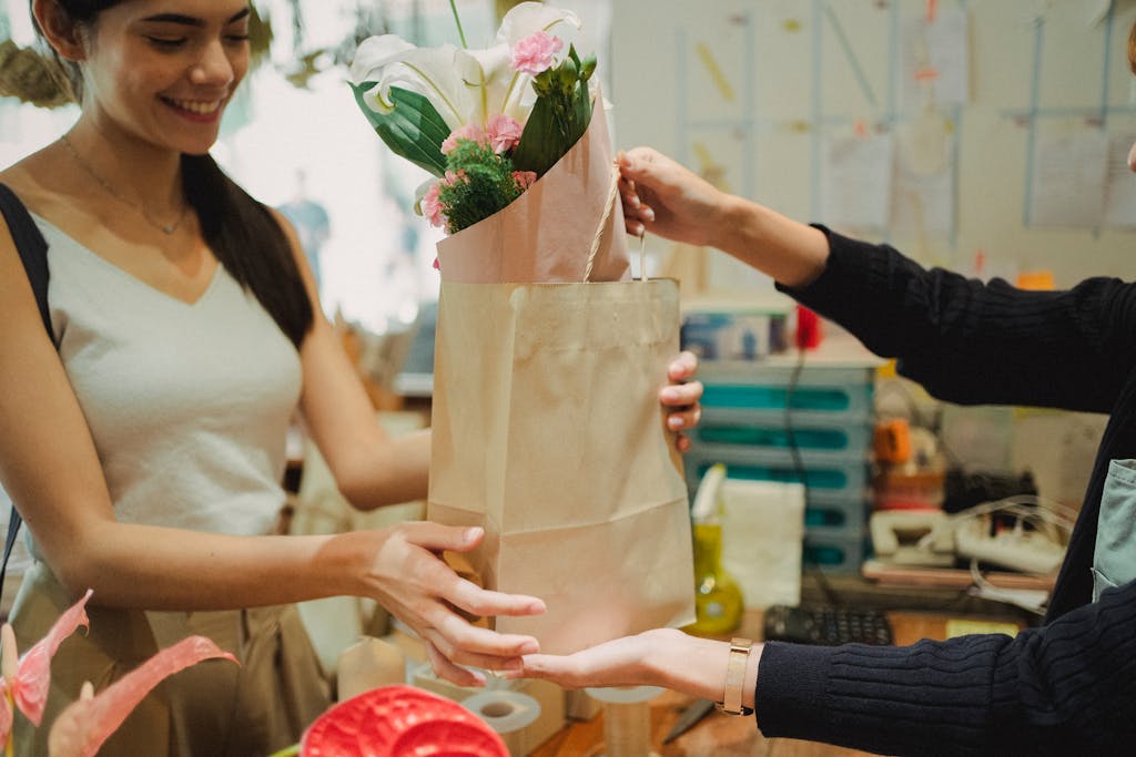 Crop florist with toothy smile passing paper bag with bouquet of flowers to faceless client in store