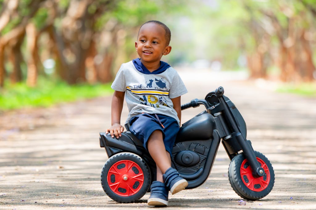 Happy child sitting on a toy scooter with legs crossed, smiling during a sunny day on a peaceful path.
