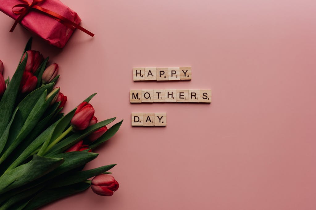Mother's Day themed image featuring tulips and gift on a pink background with letter tiles spelling 'Happy Mother's Day'.