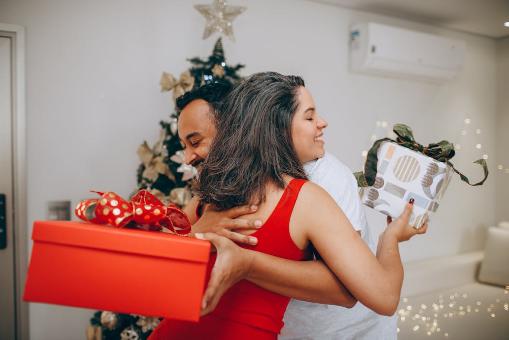 Couple hugging with wrapped presents in front of a festive Christmas tree indoors.