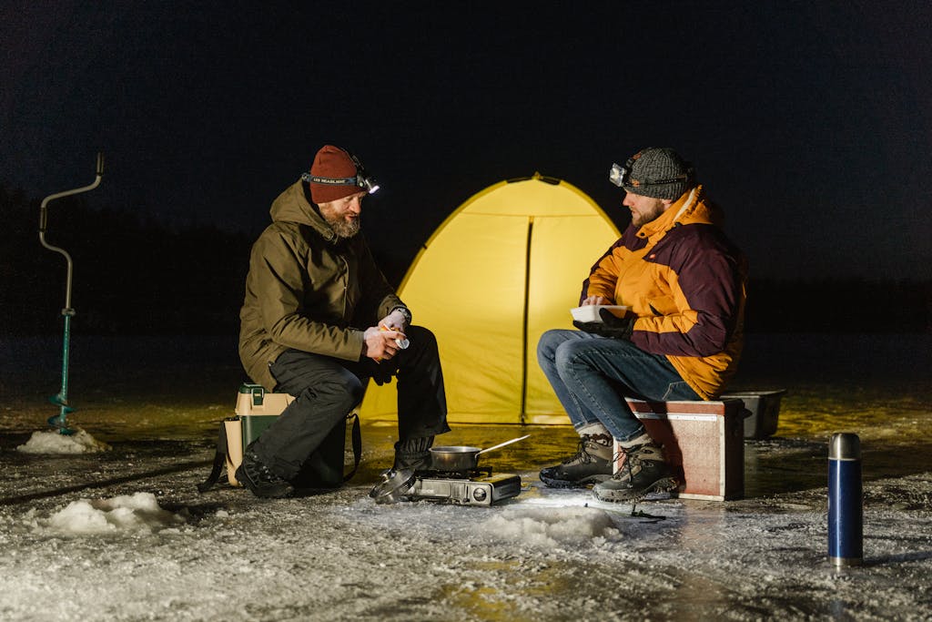 Two men ice fishing at night with a tent on a frozen lake, enjoying a warm meal.