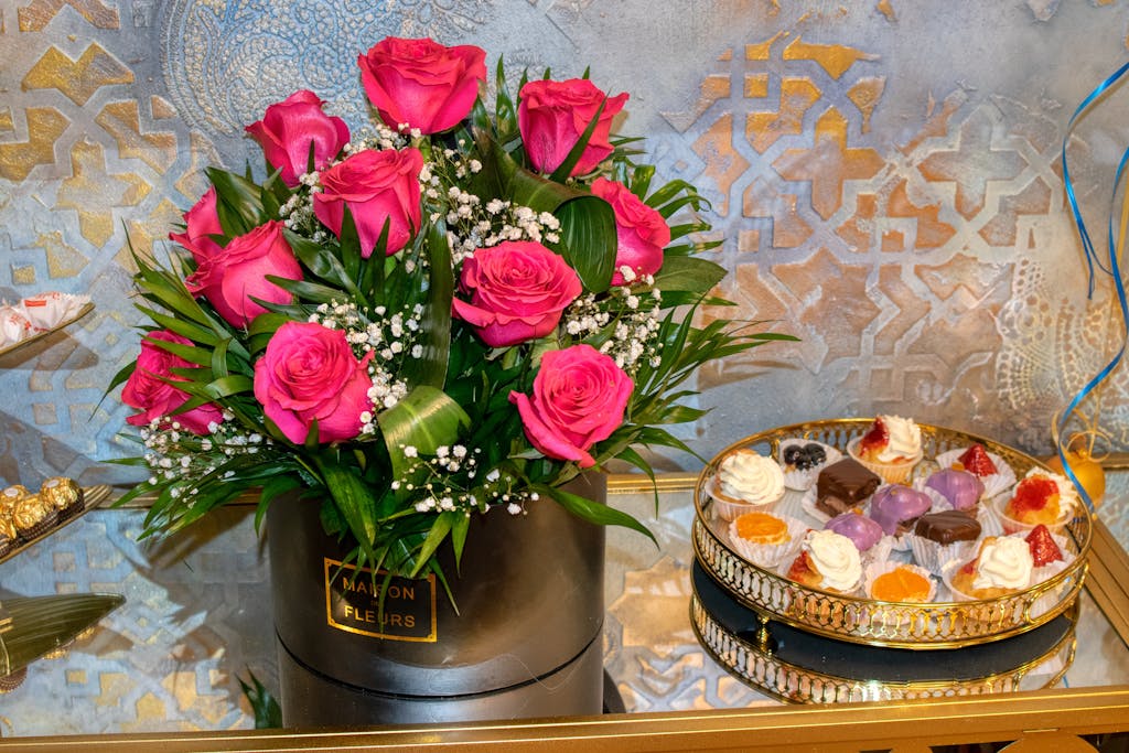 Vibrant pink roses in a chic arrangement next to a tray of assorted desserts on a decorative table.