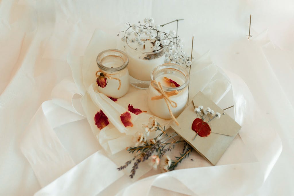 From above of twigs of flowers placed on white fabric with vintage envelope and jars with scented candles in light room