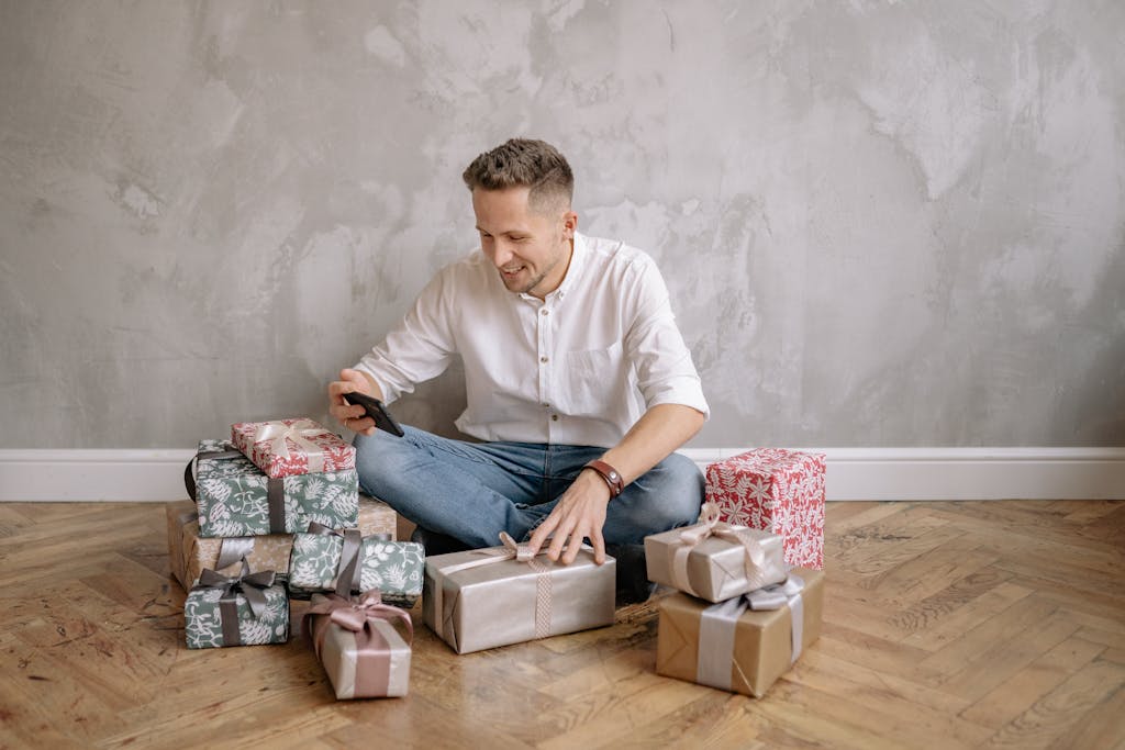 Man using smartphone while sitting on floor surrounded by wrapped gifts in a cozy room.