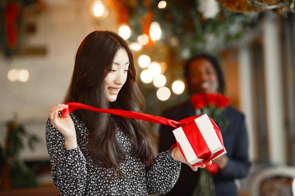 Young woman excitedly unwrapping a gift box while a person holds flowers in a warm, festive setting.