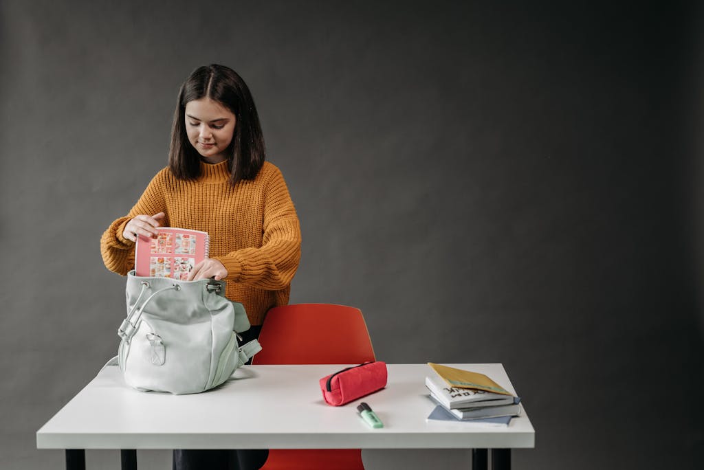 Caucasian girl organizing school supplies into her backpack at a desk.
