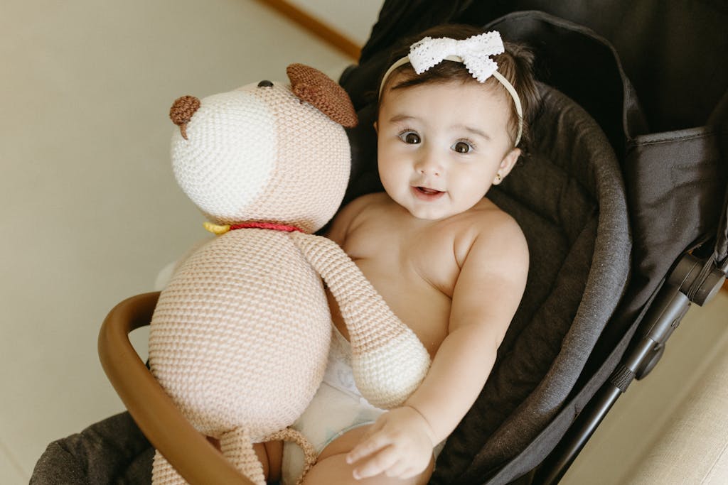 A cute baby in a stroller holding a knitted teddy bear inside a cozy indoor setting.