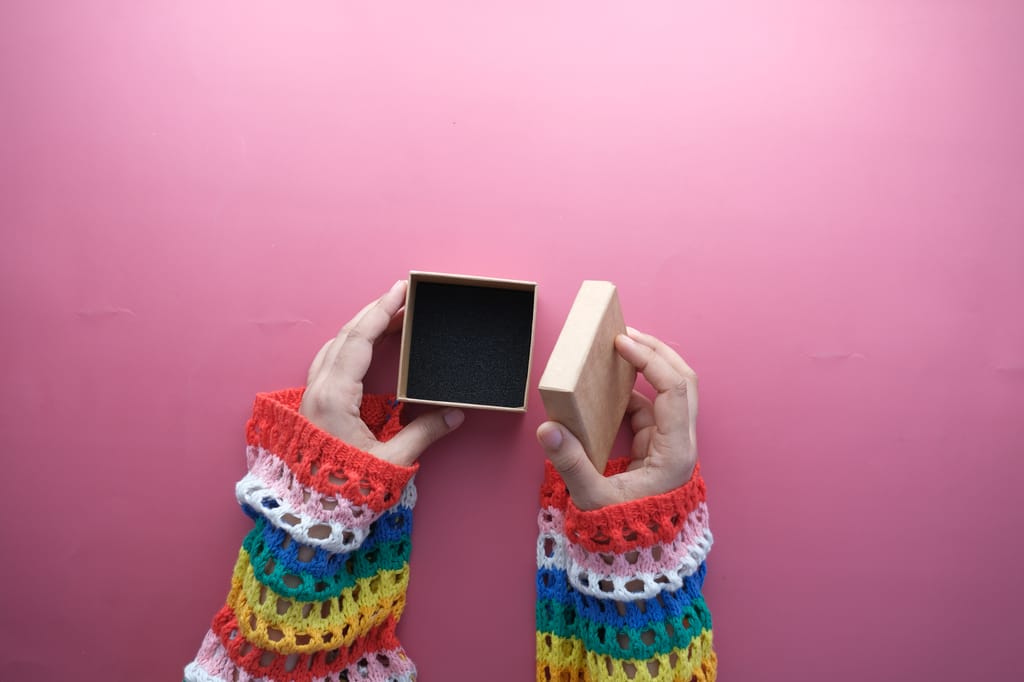 A close-up of hands in colorful sleeves opening a jewelry box on a pink background.